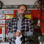Bearded mechanic standing in a garage surrounded by license plates and car parts.